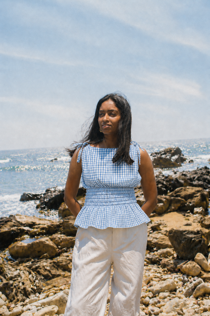A woman standing on a rocky beach wearing a light blue and white gingham sleeveless top with a peplum waist and white wide-leg pants, with the ocean and sky in the background.