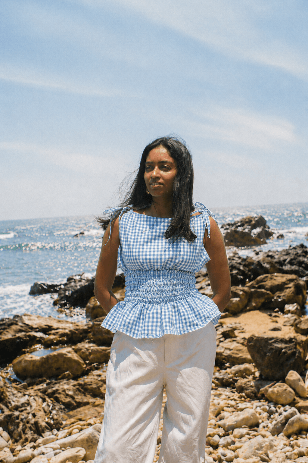 Woman standing on a rocky beach wearing a blue checkered top and white pants.