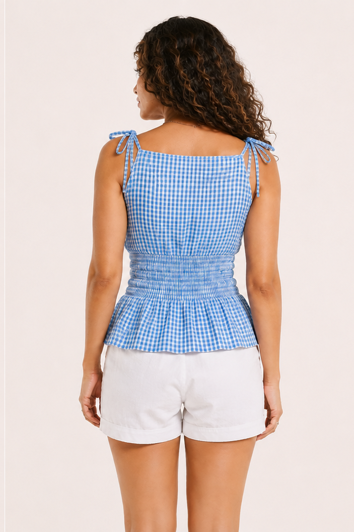 A model shown in back view wearing a light blue and white gingham peplum top with tie straps and white shorts, against a solid pale pink background.