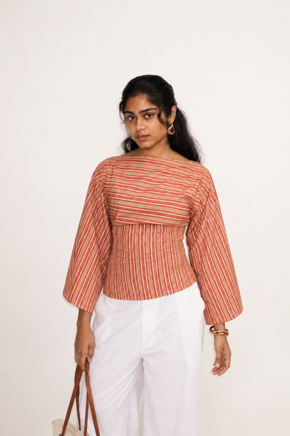 A woman modeling a cropped, off-the-shoulder top with vertical red, orange, and beige stripes, paired with white pants, against a plain white studio background.