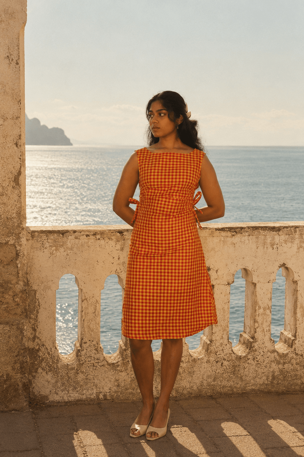 Woman in an orange checkered dress standing on a balcony with ocean view