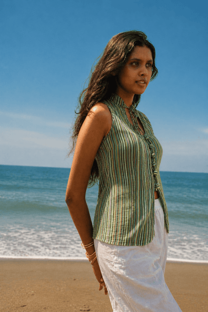 Woman in a green striped shirt and white pants standing on a beach with blue sky and ocean.