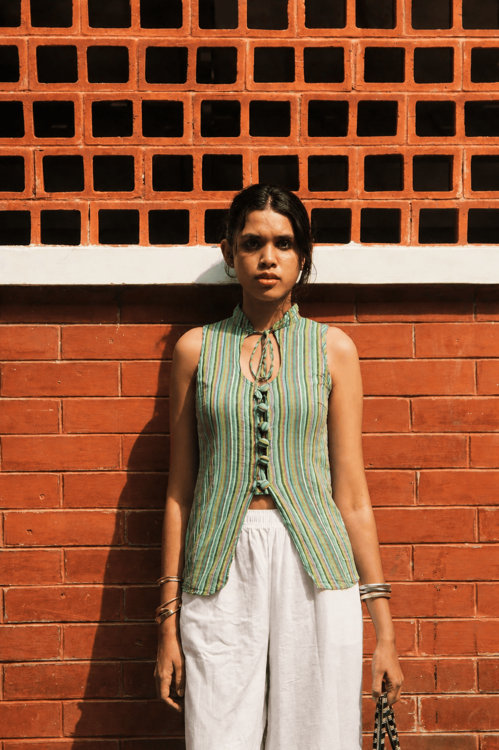Woman wearing a green sleeveless top and white pants standing against a red brick wall.