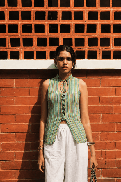 Woman wearing a green sleeveless top and white pants standing against a red brick wall.