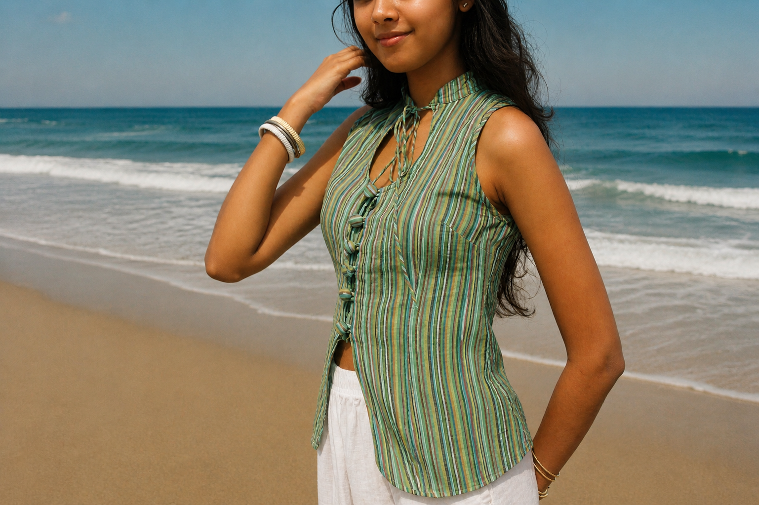 A woman standing on a sandy beach wearing a sleeveless green and yellow striped top with white pants, with the ocean in the background.