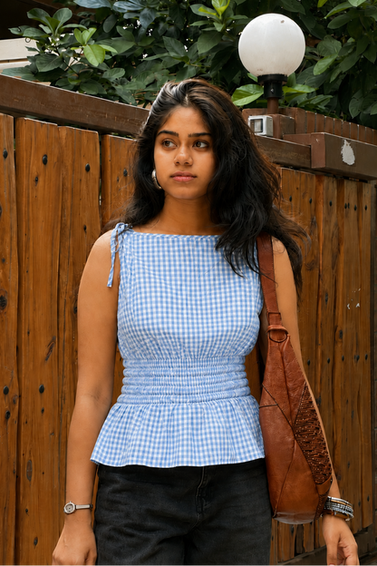 A woman wearing a light blue and white gingham check peplum top, black pants, and carrying a brown leather shoulder bag, standing in front of a wooden fence.