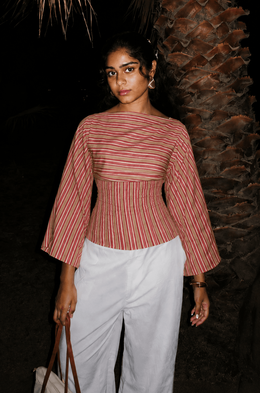 Woman in the Rose Stripe Tie-Back Top and white trousers against a dark palm backdrop