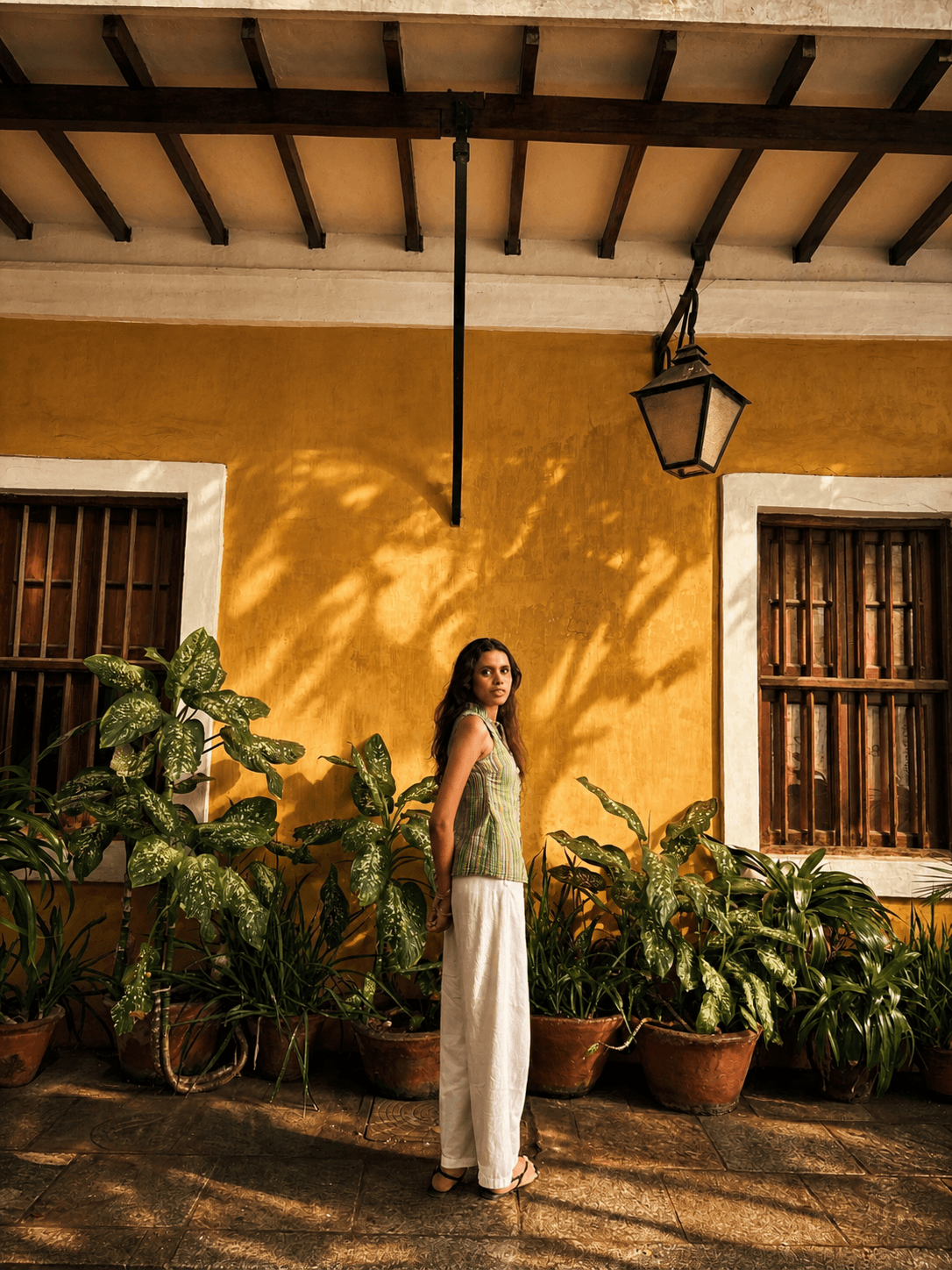 Woman in the Sundown Stripe Green Top beside a yellow colonial wall with potted plants