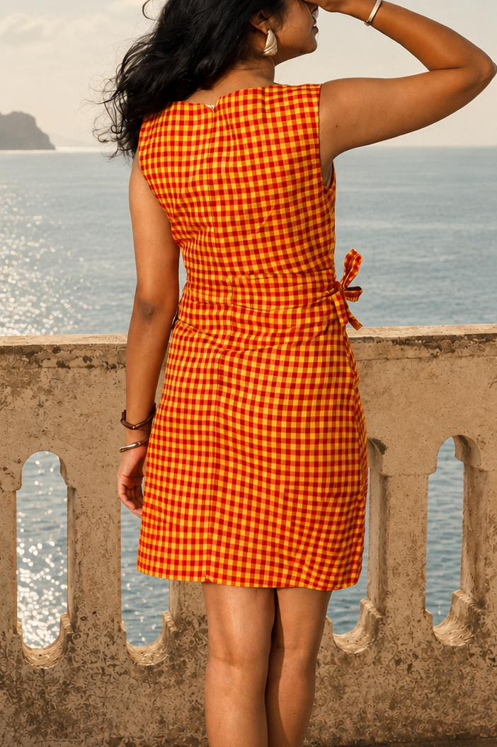 A woman stands on a stone balcony overlooking the sea, wearing a sleeveless orange and white gingham dress with a tie waist detail. The background features the ocean, distant cliffs, and a stone railing.